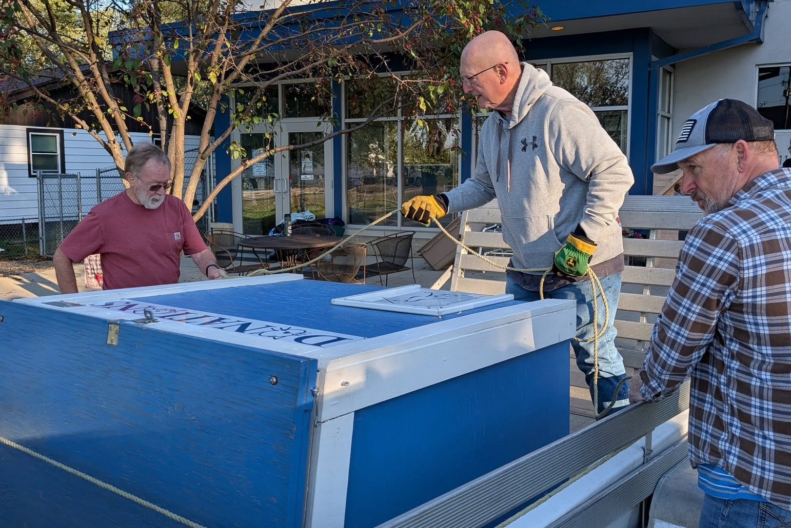 Three men loading a large blue and white box onto an aluminum trailer.