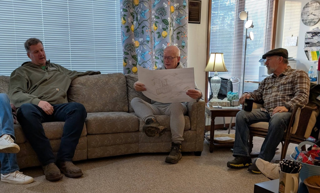 Three older men with light skin chatting in a living room.