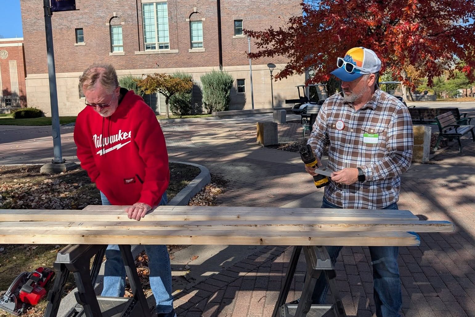 Two older men with light skin standing at sawhorses with wood.