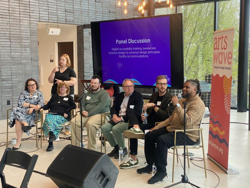 A panel discussion taking place with six people seated on a stage. One person speaks into a microphone. Behind them, a banner says "Arts Wave" and a screen displays topics like digital accessibility and design principles. An interpreter is using sign language to the side for the audience.