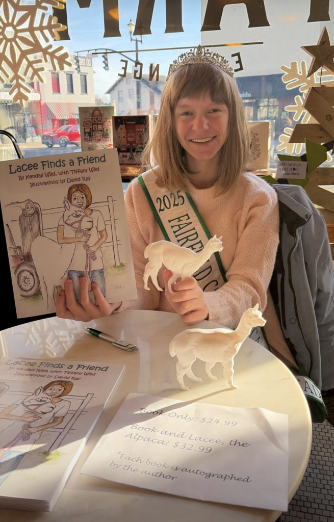 Young person in a sash and tiara, holding a children's book with an illustrated cover featuring a person who looks like them. The book is titled "Lacee Finds a Friend" and cover features a smiling person hugging an alpaca.