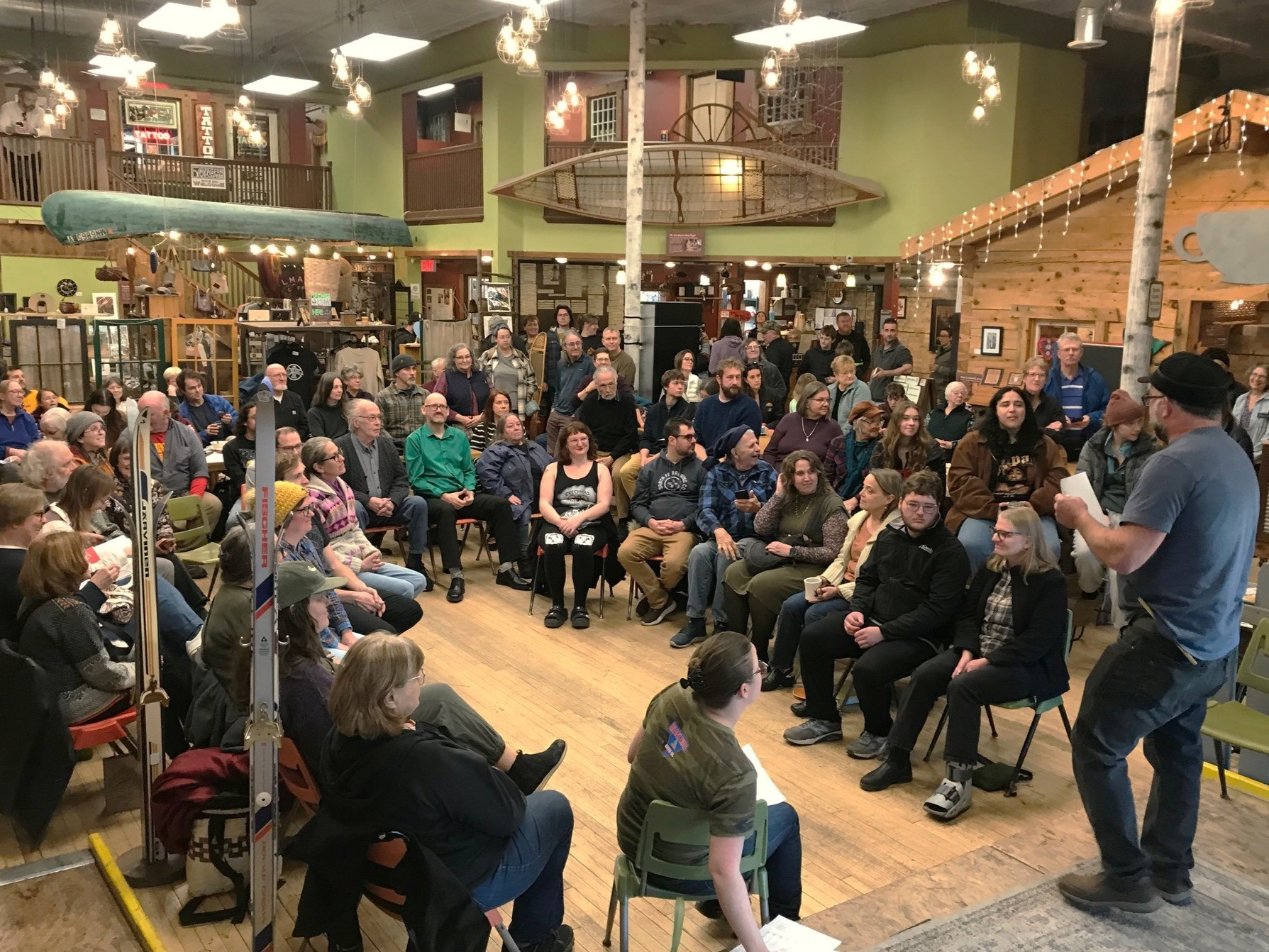 A large room holding a large, mostly sitting, crowd of people gathered in a semicircle.