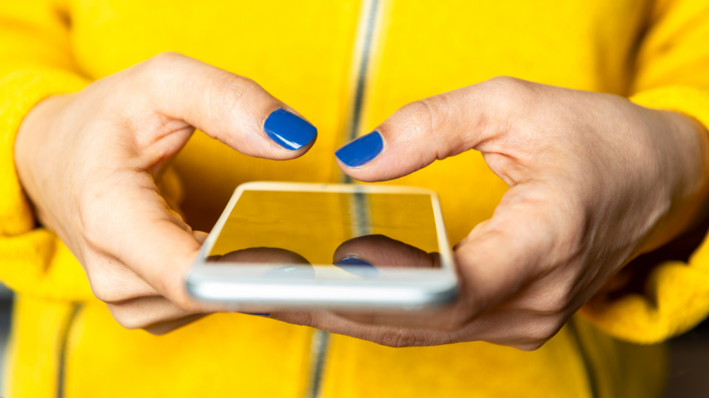 Close-up of hands holding a smartphone, with blue-painted nails and wearing a bright yellow jacket.