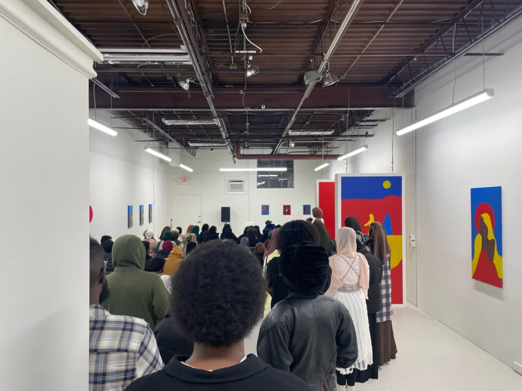 A packed gallery opening with a diverse crowd facing bold, flat-style paintings in primary colors depicting figures in desert landscapes. The white-walled industrial space is filled with standing attendees.