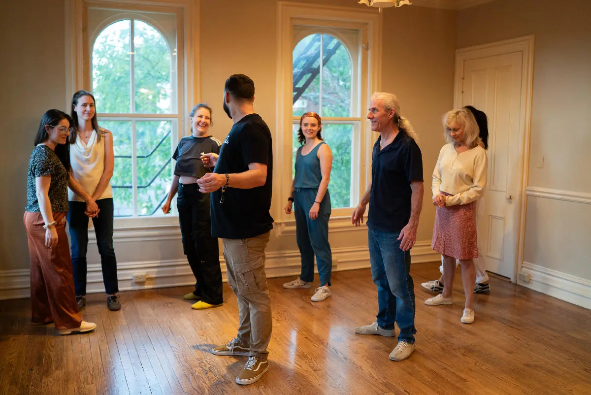 A group of people casually chat and laugh in a Dance hall with bright natural light, wooden flooring and large windows.